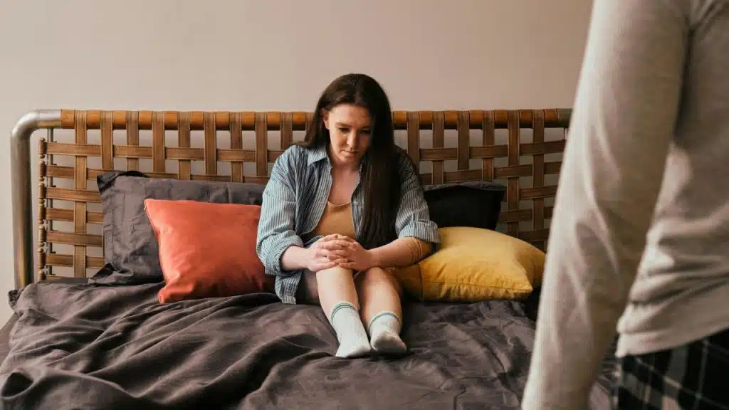 Woman sits alone on a bed with her knees clasped, looking down, facing a blurry figure.