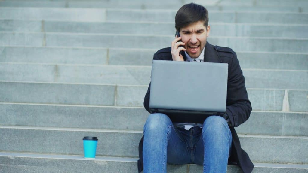 A man sits on concrete stairs, working on a laptop, talking on a phone, with a blue cup nearby.