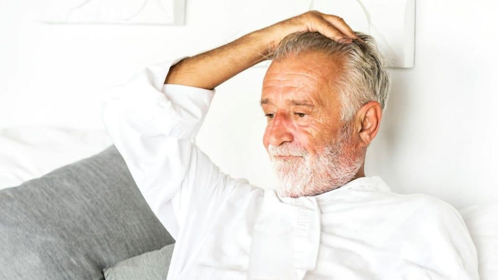 Older man with white beard and shirt touching his head, sitting on a couch.