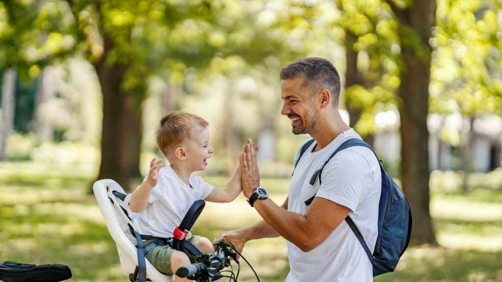 A father and son smiling and high-fiving while biking in a park.