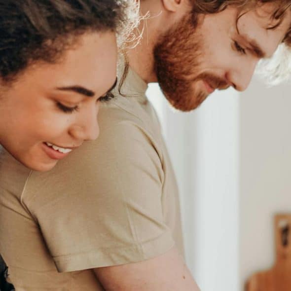 A woman smiling while hugging a man from behind in a bright kitchen.