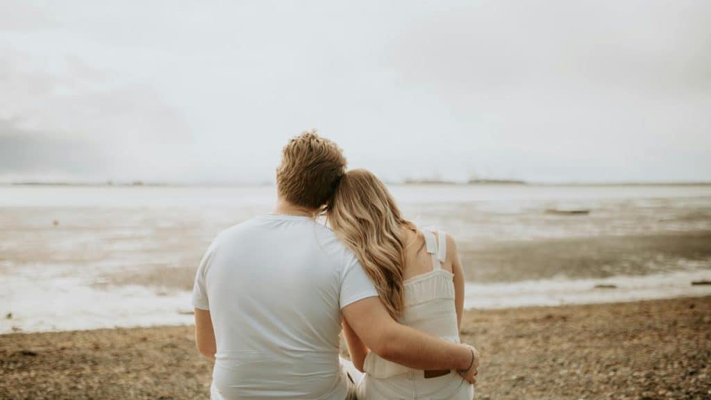 A couple sitting on a beach, with the woman resting her head on the man's shoulder.