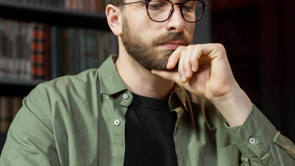 A bearded man wearing glasses sitting and thinking.