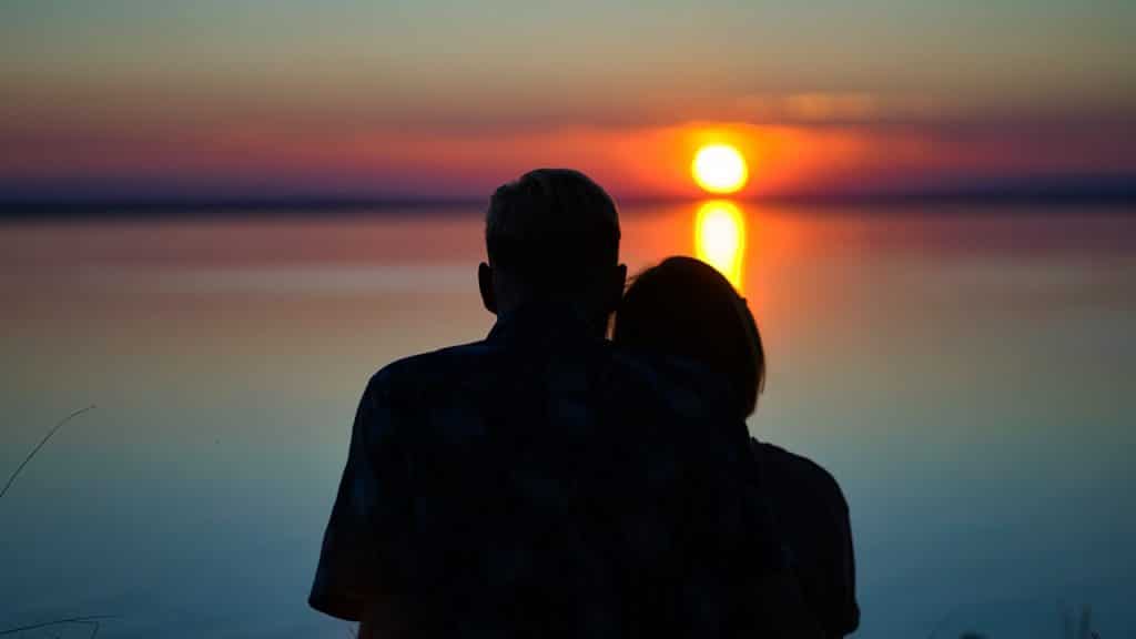 A silhouette of a couple, viewed from behind, watching a sunset over a body of water.
