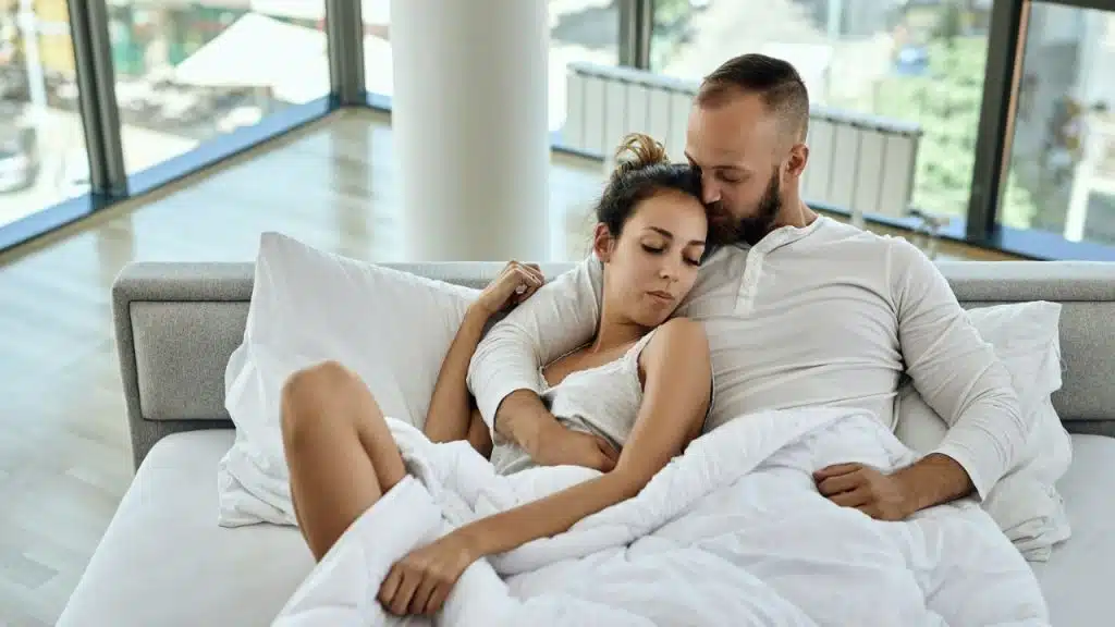 A man is embracing and kissing a woman's forehead while they are lying in bed.