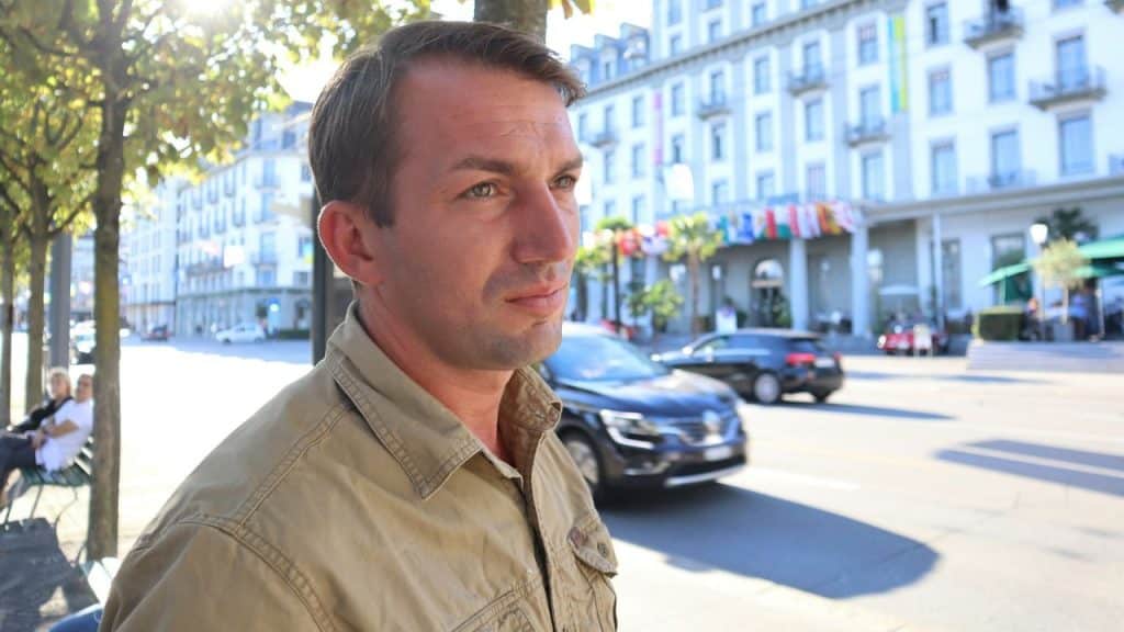 A man in a tan shirt is looking to the side on a sunny city street.