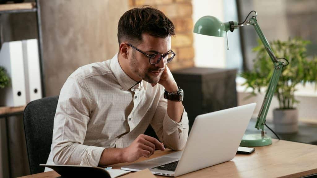 A man in glasses is intently working on a laptop at a desk with a green lamp.