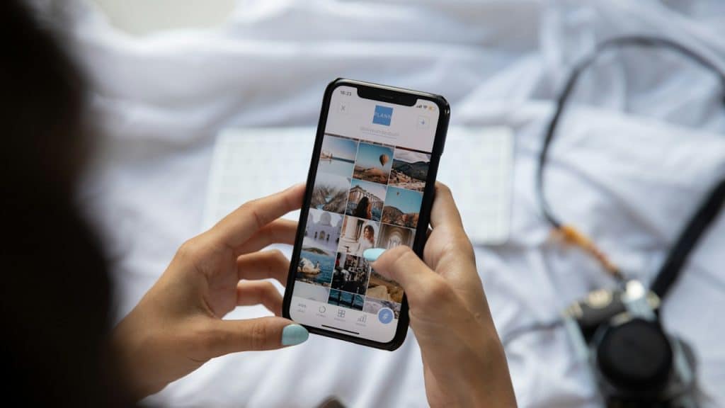 Two hands holding a smartphone displaying a grid of photos on a social media profile, resting on a white surface.