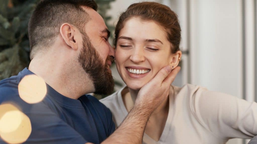 A smiling woman with her eyes closed being affectionately touched on the cheek by a bearded man.