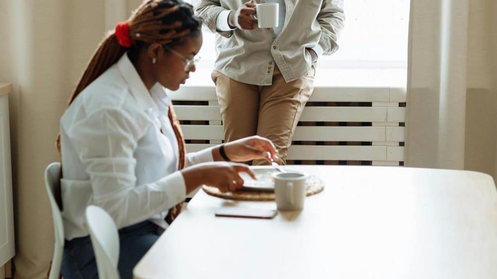 A woman using a tablet at a table while another person stands with a mug.