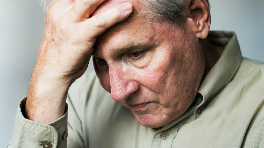 A senior man with white hair holds his head in his hand, looking troubled.