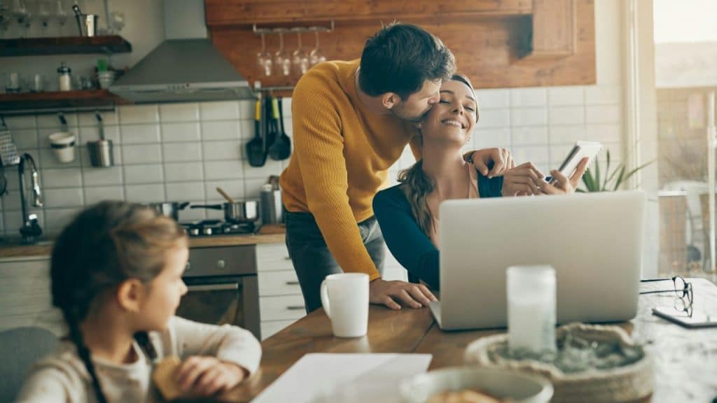 A man kisses a woman working on a laptop while a young girl sits at the table.