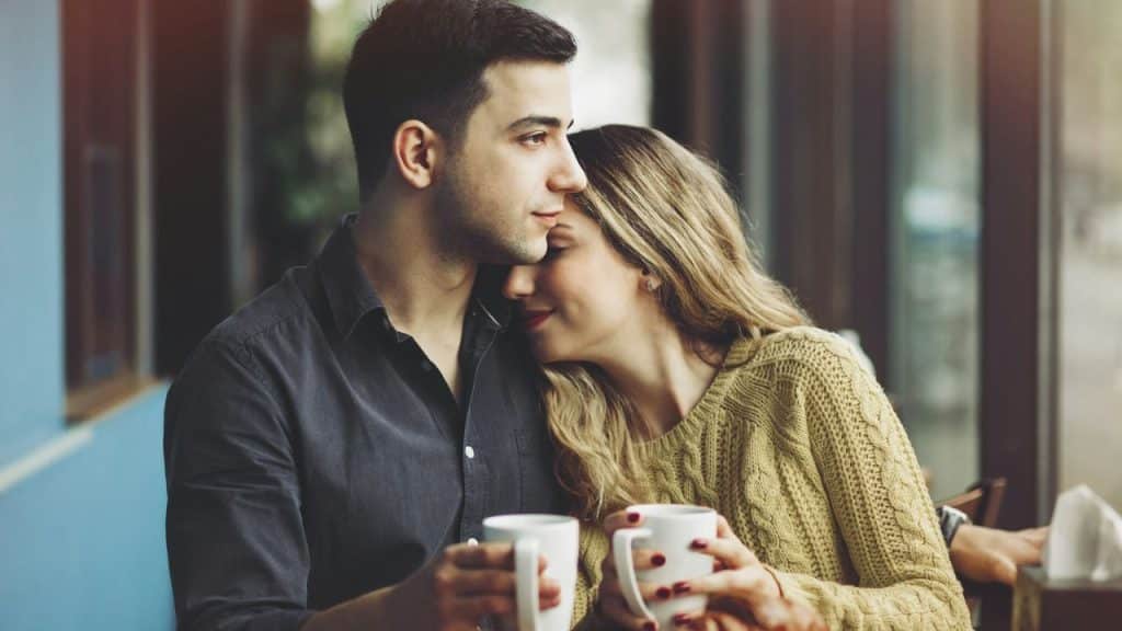 Couple seated indoors, holding coffee mugs, with the woman leaning her head on the man's shoulder.
