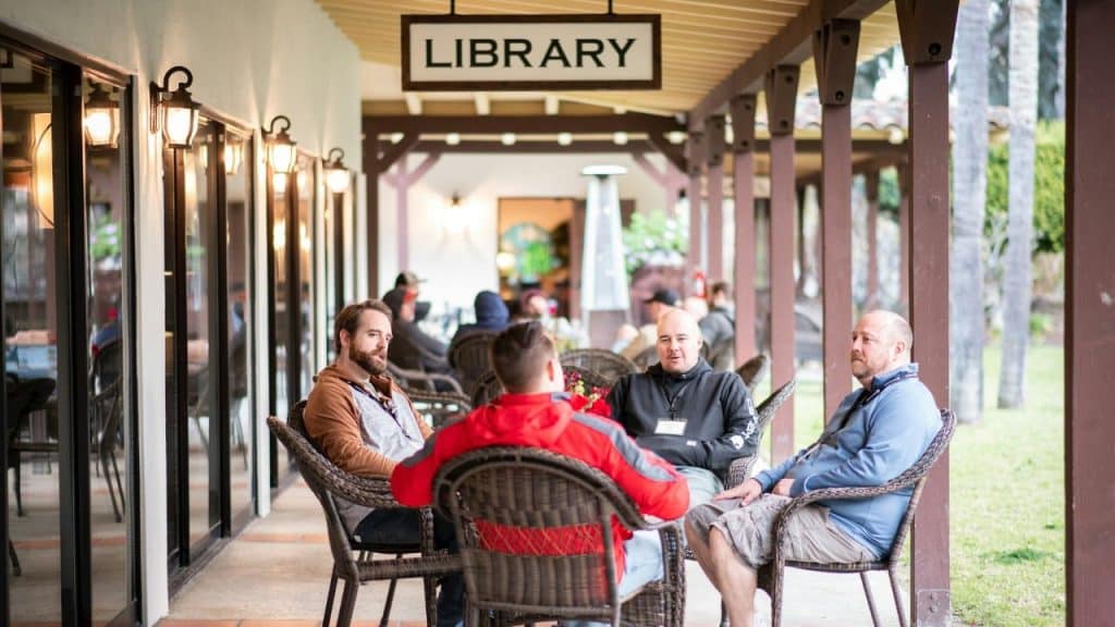A group of men are sitting and talking on a covered patio labeled "LIBRARY."