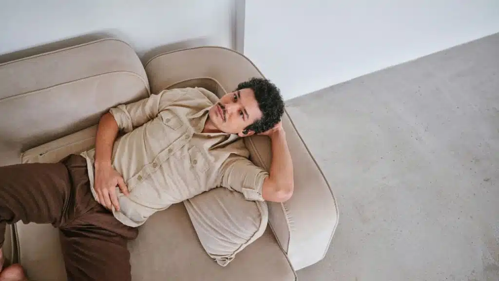A man with a mustache is lying on a light-colored couch, wearing an earbud.