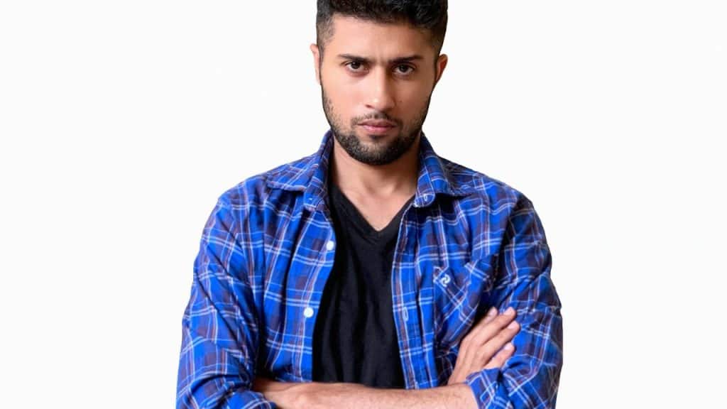Close-up of a man with a serious expression, wearing a blue plaid shirt with arms crossed.
