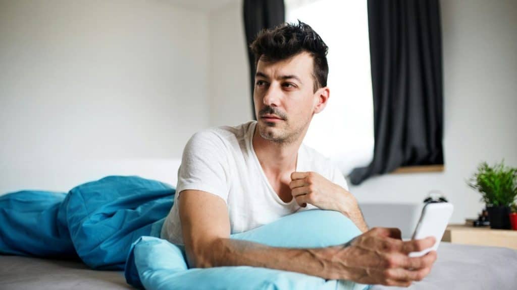 Man in a white t-shirt lying in bed under a blue blanket, holding a smartphone and looking away thoughtfully.