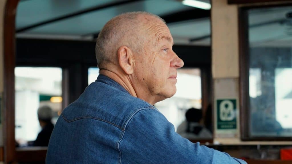 Older man with a bald head wearing a denim shirt, looking thoughtfully to the side indoors.