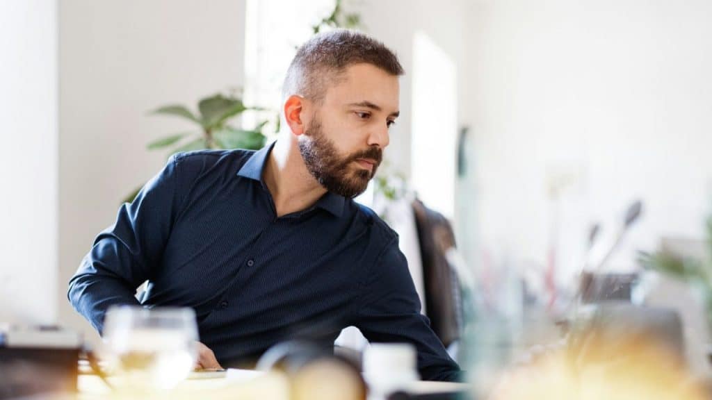 Bearded man in a dark blue shirt looking down and leaning forward in an office.