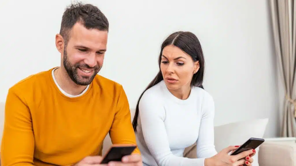 A man smiles at his phone while a woman glances at his screen with a worried expression.