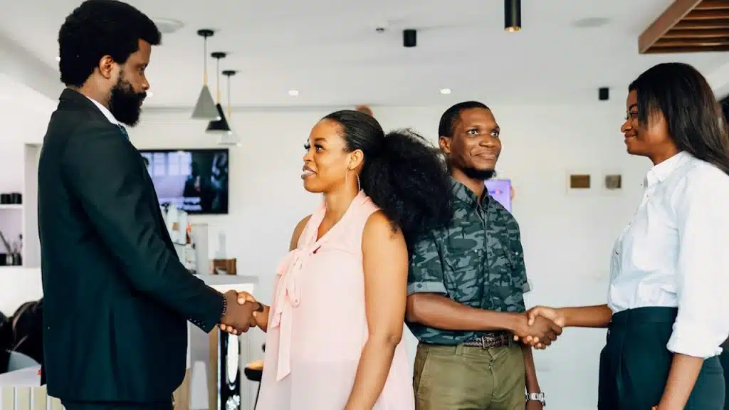A group of four business people are standing around a table shaking hands and talking.
