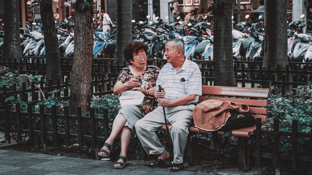 An elderly couple sitting closely on a wooden park bench in a city, talking and smiling at each other.