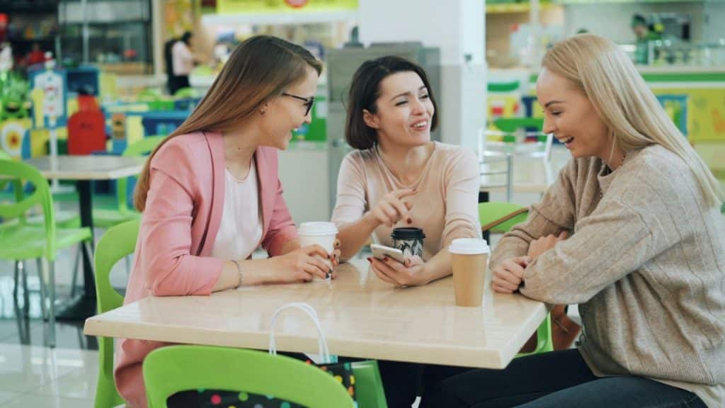 Three women sit at a table in a bright public area, smiling and talking while holding drinks.