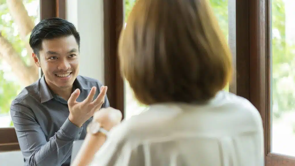 Smiling man in a grey shirt talking animatedly to a woman, facing away.
