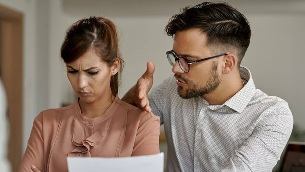 Man with glasses touches a frowning woman's shoulder while discussing papers.