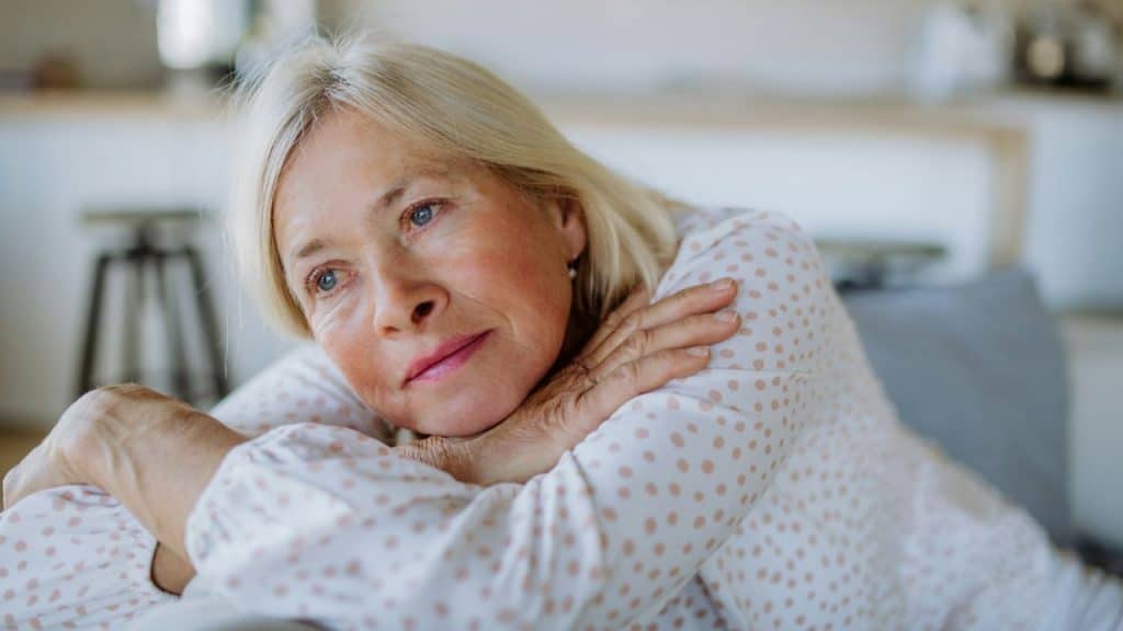 Older woman with blonde hair rests her head on her folded arms, looking away.