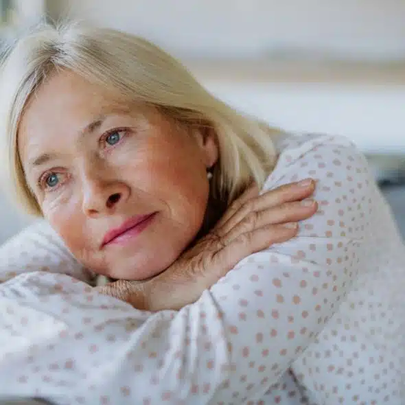 Older woman with blonde hair rests her head on her folded arms, looking away.