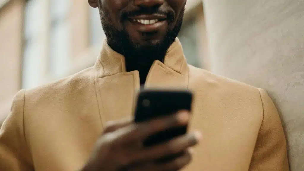 A man wearing a tan coat smiling while looking at his phone.