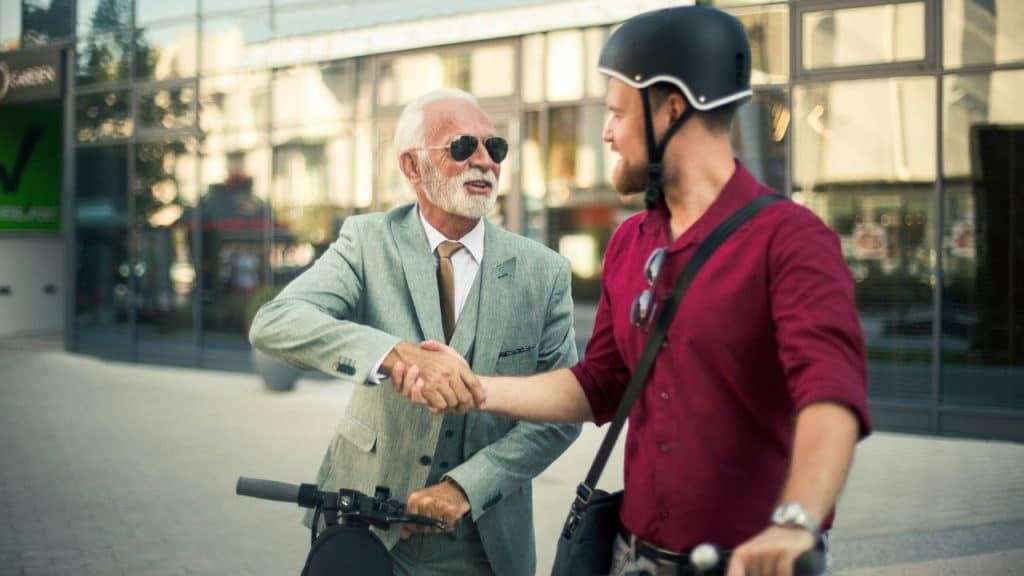 Two men, one in a suit and one on a scooter, shake hands outside a building.