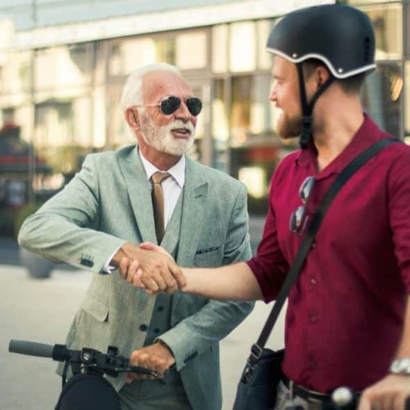 Two men, one in a suit and one on a scooter, shake hands outside a building.