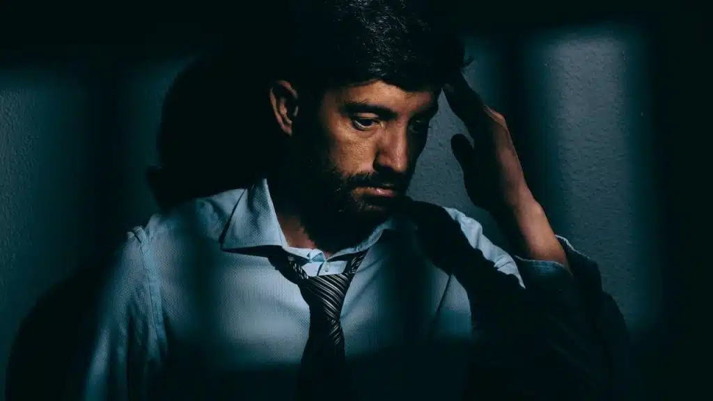 Man with beard and tie, looking down with hand on head in a dark setting.
