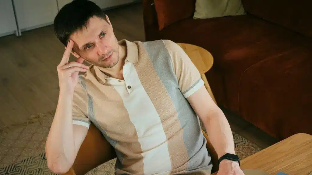 Man in a striped polo shirt sitting in a chair with his hand on his temple.