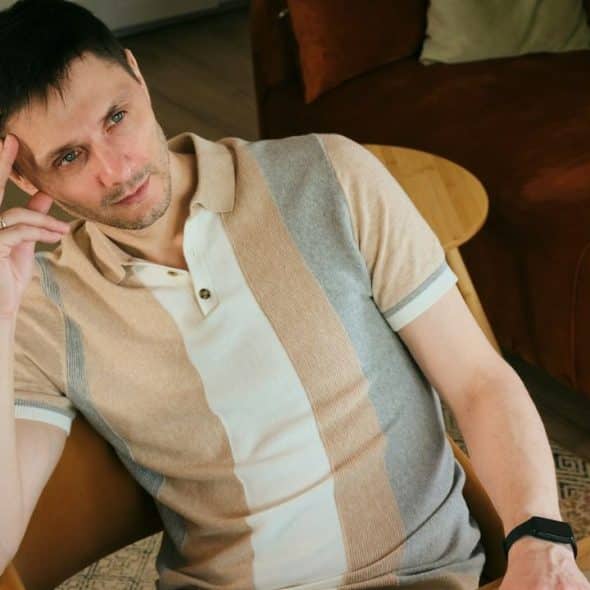 Man in a striped polo shirt sitting in a chair with his hand on his temple.