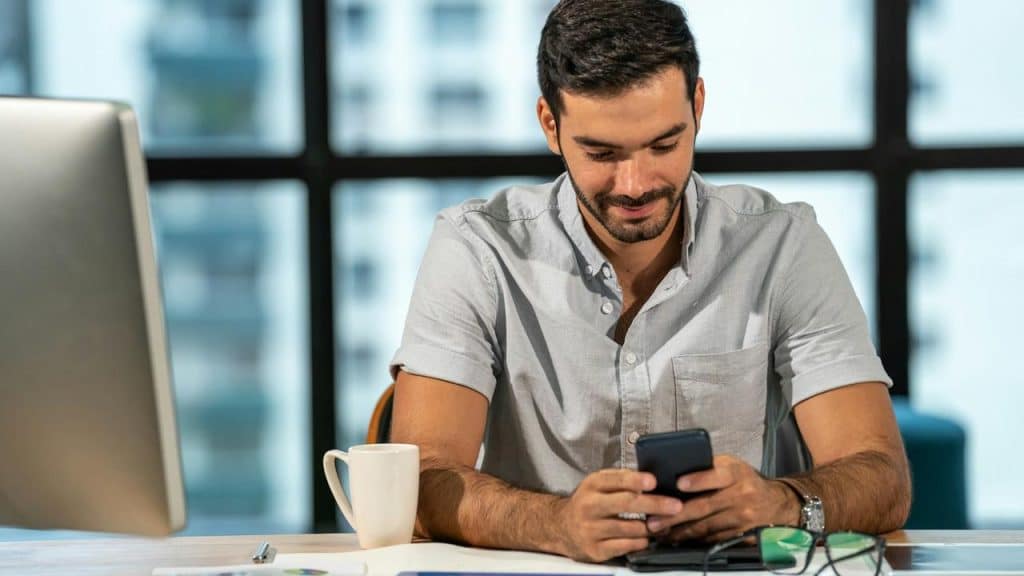 A man sitting at a desk smiling while looking at his smartphone.