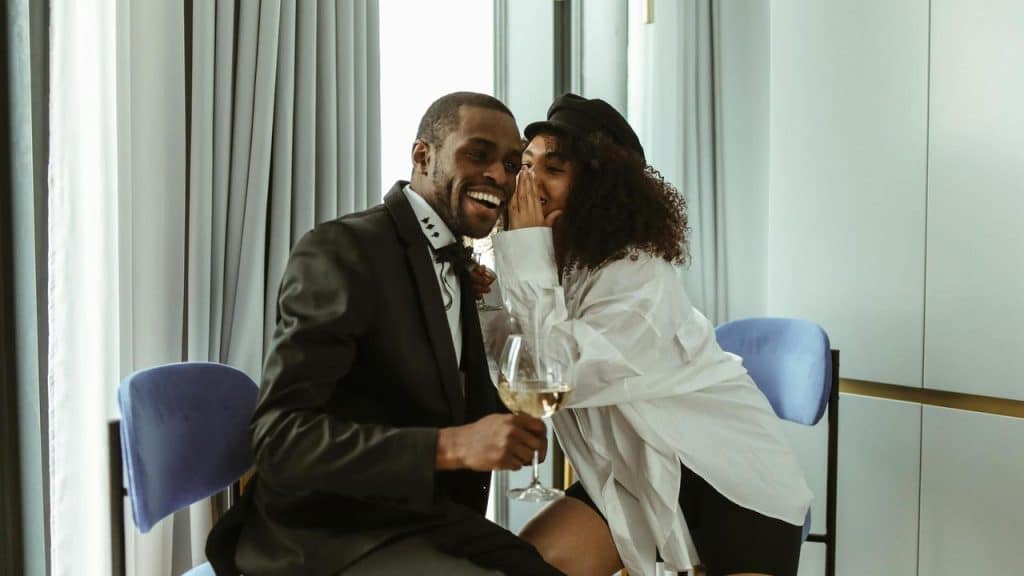 A woman whispering in a man’s ear while they laugh and hold a glass of wine.