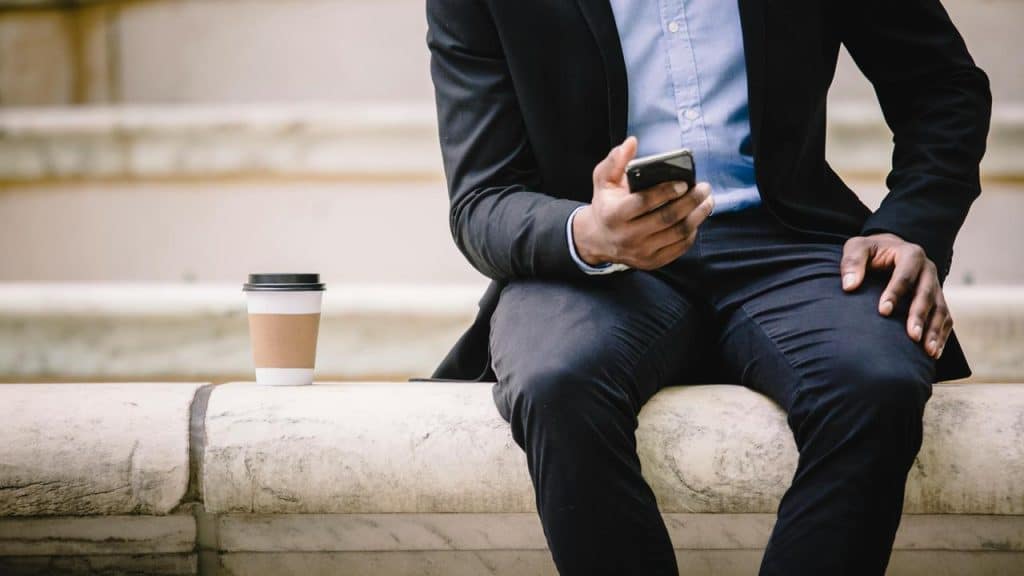 A person in business attire sitting on a ledge, holding a phone with a coffee cup nearby.