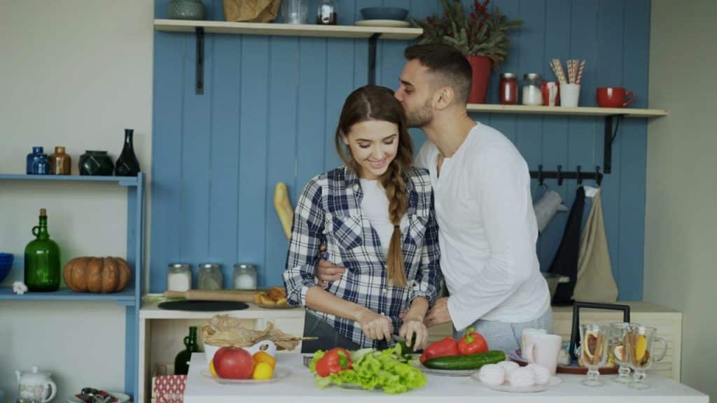 Smiling couple cooking in a kitchen, man kisses woman's forehead.