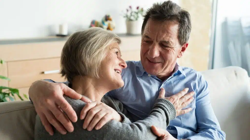A loving, close-up shot of a smiling, middle-aged couple embracing on a couch, looking warmly into each other's eyes.