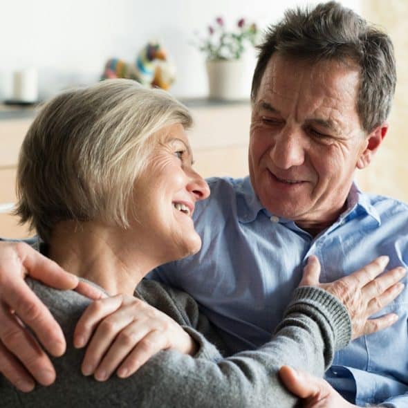 A loving, close-up shot of a smiling, middle-aged couple embracing on a couch, looking warmly into each other's eyes.