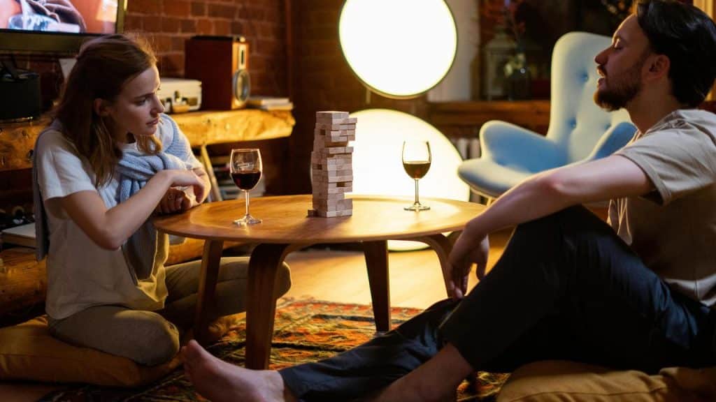 A pair of people sitting on the floor playing a block stacking game with glasses of wine.
