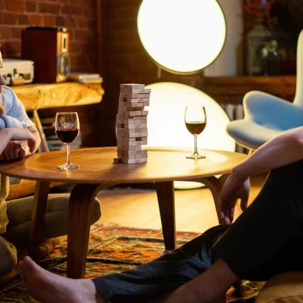 A pair of people sitting on the floor playing a block stacking game with glasses of wine.