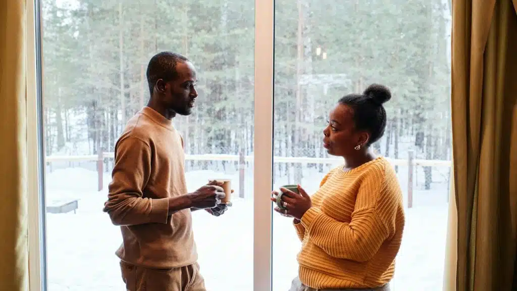 A couple holding coffee mugs are talking by a window.