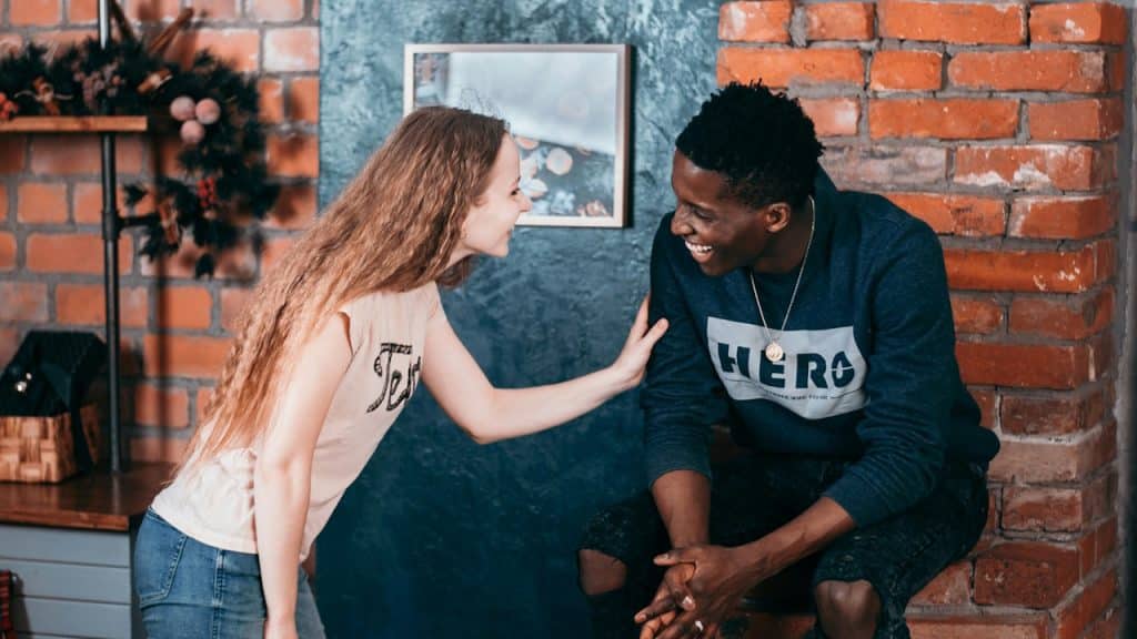 A smiling young woman with long wavy hair leaning in and touching the arm of a laughing young man.