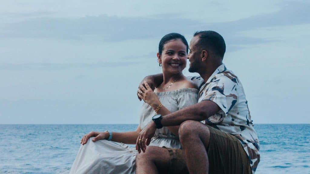 A couple sitting by the ocean, smiling at each other with calm water in the background.