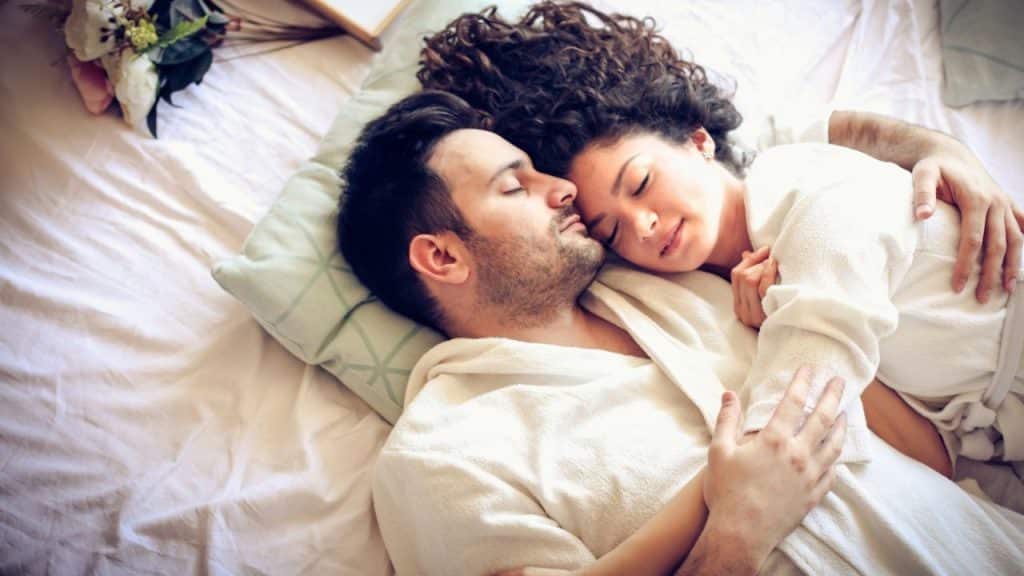 A man and a woman in white robes are sleeping embraced in a bed.