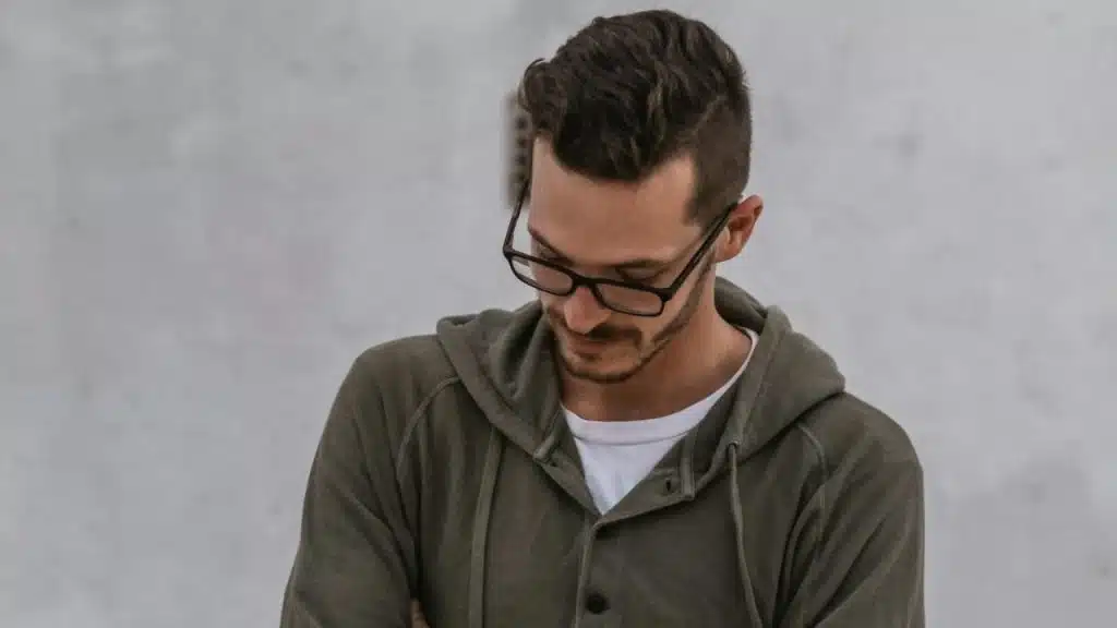 A man in a green hoodie and glasses is looking down with a somber expression.